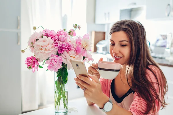 Женщина Пьет Кофе Используя Телефон Кухне Украшенный Розовыми Пионами Девочка — стоковое фото