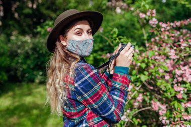 Kadın fotoğrafçı, yaz bahçesinde koronavirüs koruyucu maske takarak fotoğraf çekiyor. Parkta yürüyen serbest yazar çiçeklerin açtığı çalıları filme alıyor.