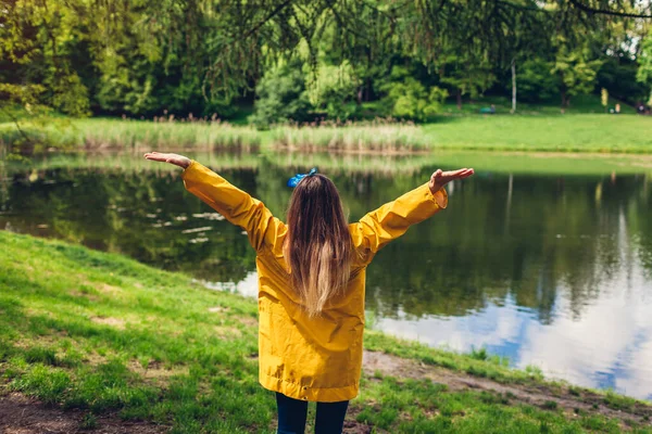 Happy woman in yellow raincoat raising arms feeling free walking in summer park. Girl admires lake landscape view. Harmony and balance with nature