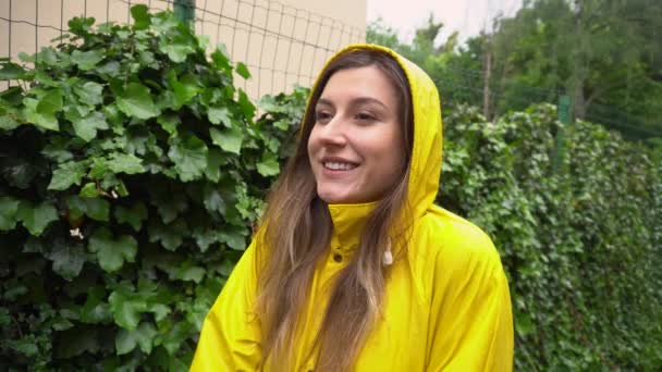 屋外で夏の雨を楽しむ黄色のレインコートで陽気な若い女性 女の子笑みを浮かべて腕を上げる感じ幸せな霧雨の下 — ストック動画