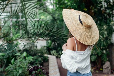 Şık yaz aksesuarları. Hasır şapkalı, tropik egzotik serada yürüyen genç ve modaya uygun bir kadının portresi..