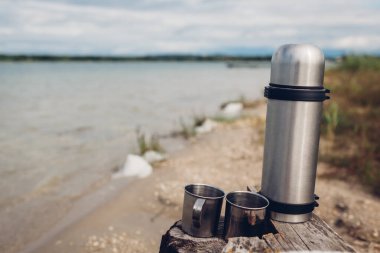 Yaz gölünün yanında sıcak çay ve metal bardaklı termos vakum şişesi. Açık havada içkilerle ısınıyorum. Kamp, seyahat ve turizm konsepti