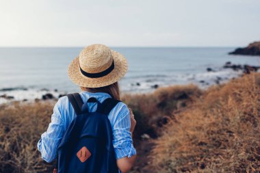 Sırt çantalı bir kadın gezgin deniz kıyısında yürüyor manzaranın tadını çıkarıyor. Turizm, seyahat. Yaz tatili