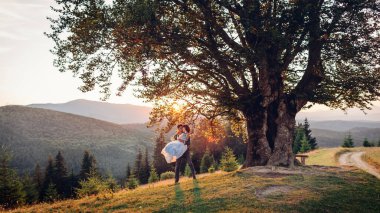 Yeni evlilere bayılan çiftler gün batımında dağlarda birbirlerine sarılıyorlar. Damat gelini büyük ağacın altında tutuyor. Yazın düğün Karpatlar.