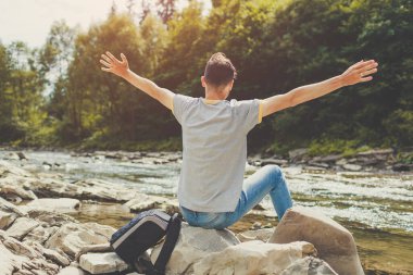 Dağ nehri kıyısındaki kayalıklarda oturup doğal manzaranın tadını çıkaran adam. Gezgin sırt çantalı mutlu kollarını kaldırıyor. Yaz gezisi