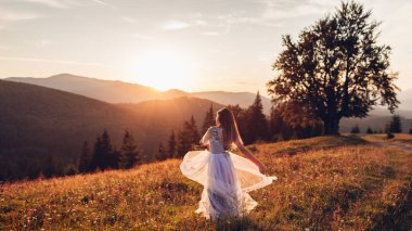 Güzel gelin, gün batımında Carpathian dağlarında mavi gelinlik içinde dans ediyor. Kadın elbise tutan manzarayı seviyor. Arka plan