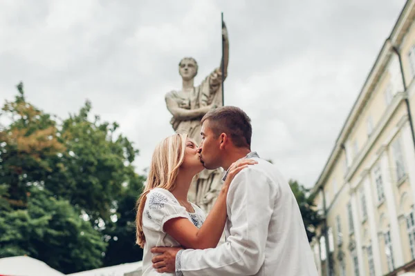 Young couple in love kissing in old Lviv city by fountain statue wearing traditional ukrainian shirts. Man and woman walk on square