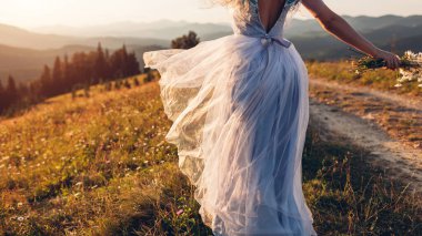 Gelin, gün batımında dağlarda buket tutan, mavi, sırt dekolteli bir elbise giyiyor. Etek kumaşı rüzgarda sallanıyor. Arka plan