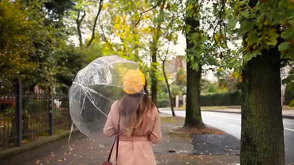 黄色の葉を保持する雨の間に透明な傘の下で秋の街の通りを歩く保護マスクのスタイリッシュな若い女性 コロナウイルス Covid 19ファッション — ストック動画