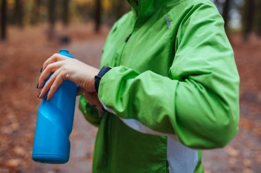 Koşucu, sonbahar parkındaki eğitim sırasında elektronik kol saatinin nabzını kontrol ediyor. Kadın su şişesiyle dinleniyor. Spor aletleri