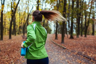 Sonbahar parkında koşucu eğitimi. Elinde su şişesiyle koşan bir kadın. Aktif sağlıklı yaşam tarzı. Antrenman yapıyorum. Sonbahar aktiviteleri. Spor.