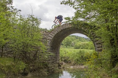 Crossing an old narrow arch bridge by mountain bike clipart