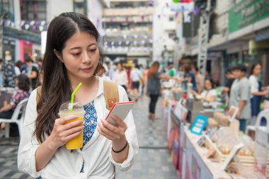Asyalı kadın mango suyu bir fincan tutarak ve yolun yanında ürünleri arıyorum. O resim çekmek için cep telefonu kullanımı.