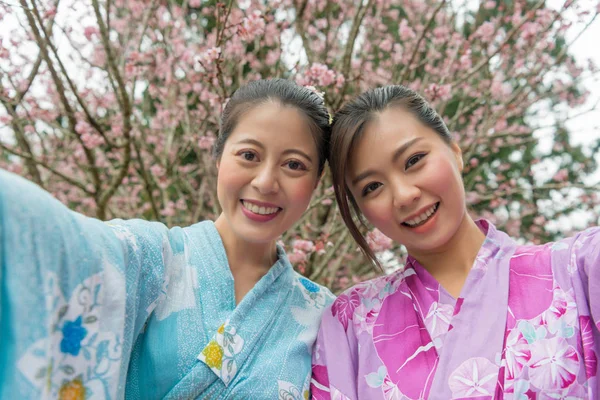 İki Asyalı kadın yan yana durmak ve bir selfie birlikte yanında kiraz ağacı almaya kollarını açtı.