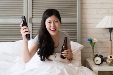 Asyalı kadın uzaktan kumanda ve bira şişesi holding elleri ve memnun mutlu biten. Arsa tablo açık.