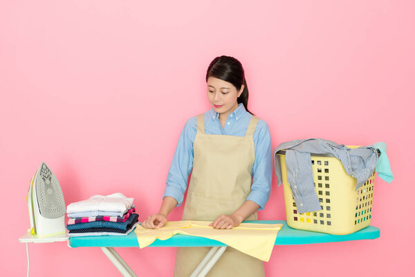 charming Japanese woman checking clean clothing on ironing board and doing laundry work on pink background
