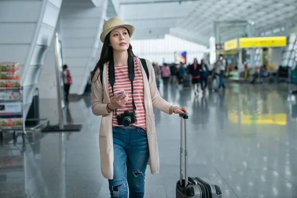 female searching for something while walking.  holding her phone in the hand. by the background of crowds in the airport.