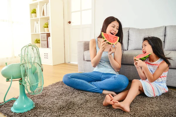 mom and kid eating watermelon happily. sitting and looking at each other pleasantly. cooling down their body with an electric fan.