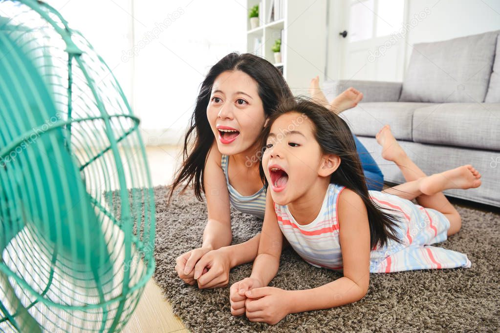 mother and child lying on the mat cheerfully. blowing their faces with electric fan.  laughing happily together.