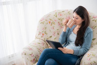 Genç ve güzel kadın oturma odasında kanepede oturan tablet kullanırken ona çay içme.