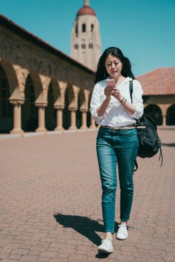 beautiful attractive asian woman browsing on a smartphone while passing through the campus. clipart