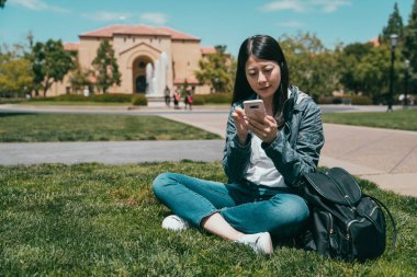 asian international student checking on smartphone and sitting on the grass in the campus. clipart