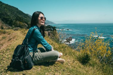 Uçurumdaki bitkilerle kaplı ve bir yere mesafe içine bakarak oturan güzel kadın.