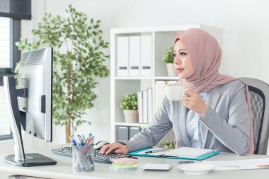Ofis masasında oturan ve bir fincan kahve tutan başarılı Müslüman hanım. Sabah bir fincan kahve yudumlarken bilgisayarda çalışarak kendine güvenen bir iş kadını portresi.