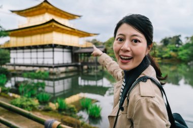 pretty female traveler pointing to the Kinkakuji temple face to camera and smiling. Sightseeing in Kyoto in Japan. backpacker lifestyle. clipart