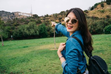 Hollywood yazısı, kamera gülen yüze işaret neşeli kadın turist. bahar tatili içinde Bayan güneş gözlüğü. Güneş ışığı ve Amerika'da yeşil doğa zevk.