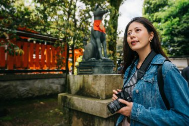 köpek heykeli önünde duran ve gökyüzüne bakarak Asya fotoğrafçı. Genç kız hobi SLR kamera ile fotoğraf çekmek. din seyahat Turizm Japonya.