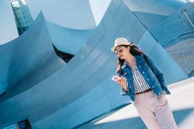 Zarif Bayan Walt Disney Konser salonu onun elinde cep telefonu ile dolaşıyor. Los Angeles'ta ziyaret için popüler gezi yer. Genç Turizm Online Tur Rehberi smartphone kullanarak.