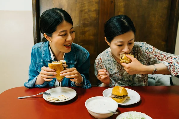 Hungry Ladies Sitting Red Table Joyfully Eating Hamburgers Asian Sisters — Stock Photo, Image