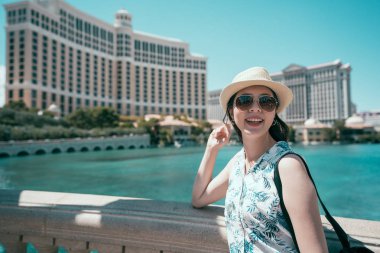Neşeli Bayan turist gülümseyen ve küpeşte üzerinde güvenerek ve su gösteri için bekleyen şapkalı. mavi su birikintisi ve güneşli gün arka planda Las Vegas uzun boylu otelin. ABD kavram kendini rehberli gezi.