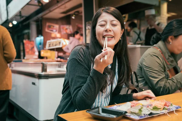 快乐的女人坐在传统市场上用筷子吃饭的摊贩 美丽的游客尝试生鱼片 享受大阪旅游概念 — 图库照片