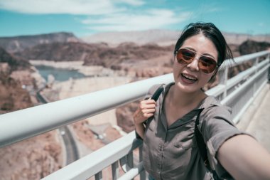 Güneş gözlüklü genç Asyalı gezgin Hoover baraj ziyaretinde kendi portresini çekiyor. Amerika 'nın güneşli doğasında zarif turistler mutlu yolculuklar. Güzel kız kameraya gülümsüyor..