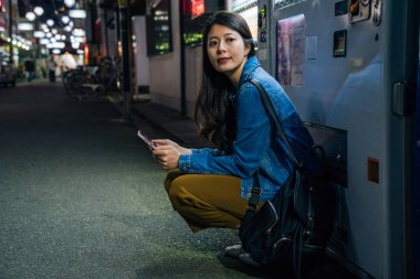 Japon kadın çömelmek tarafından otomat arkadaşımı bekliyorum. Zarif Bayan cep telefonu iletişim app açık gece şehirde kullanarak. Gezgin online harita Osaka'da Hotel'e yol bulma.