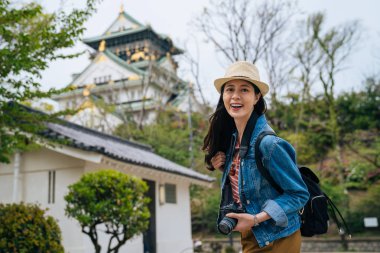 Genç kız turist yüz kamera SLR tutan gülümseyerek kamera. profesyonel fotoğrafçı her zaman onun digicam ile seyahat. Bayan gezgin güneşli yeşil ağaçlar ile Osaka'dan himeji Kalesi ziyaret.