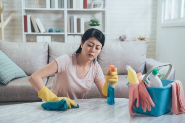 Güzel Asyalı kadın evde rahat güneş ışığı oturma odasında onun ev ev işleri yaparak. Genç Ev kadını elinde mavi bez ve koruyucu eldiven tarafından tablo silme. Kanepenin önünde katta oturan Bayan.