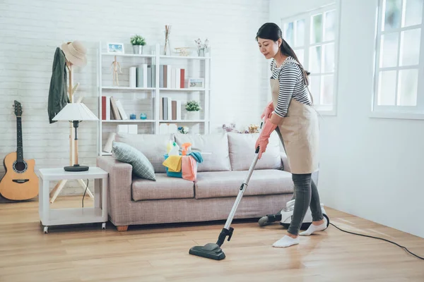 asian lady doing house chores in apron. young housewife using vacuum cleaner cleaning the wooden floor in the living room. happy housekeeper doing housework at home with attractive smile on face.