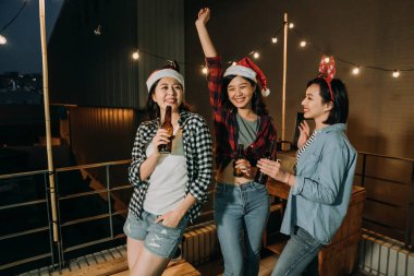 Üç Asya bayanlar çatıda karanlık gece Noel arifesi kutluyor. Barbekü partisi kadın sevinçle holding bira dans santa şapka ile. Genç kız arkadaşlar ışık asılı balkonda eğleniyor.
