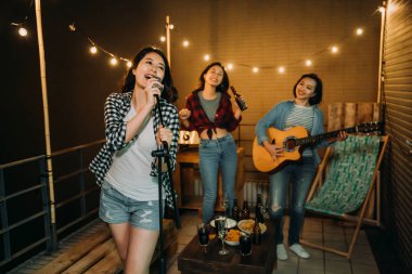 band playing music practicing songs on roof. young girls love music enjoy in karaoke party outdoor on rooftop at night. food and alcohol drinks on table beside comfortable desk chair. clipart