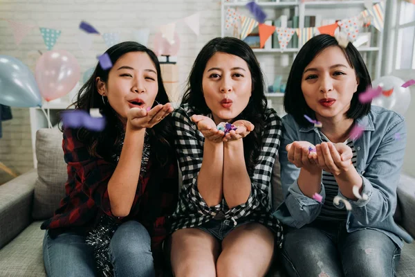 Asian cute ladies face camera cheerfully blowing colorful confetti. young girls sitting relaxing on sofa in decorated living room at home. house party full of balloons friends having fun celebrating.