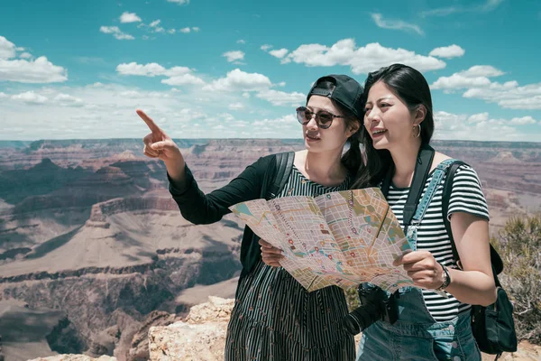 two sisters self guided tour travel together in grand canyon north rim. young girlfriends holding paper map pointing to the top of the desert standing on skywalk in national park. asian lady tourism.