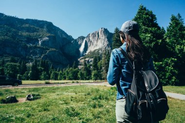 Arkadan Görünüş uzun yürüyüşe çıkan kimse kadının yosemite Milli Parkı güzel dağ görünümünde gezi çimenlerin üzerinde yürüyüş şapkalı. Genç kız backpacker tadını güneşli yolu ormandaki yakınındaki doğa ayakta