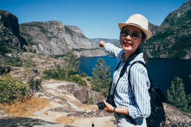 Genç kız Asya backpacker yosemite Milli Parkı ziyaret edin. zarif kadın gezgin işaret mavi gökyüzü ve temiz Gölü yüz kamera gülümseyen güneş gözlüklü. seyahat san franisco yaşam tarzı kavramı.