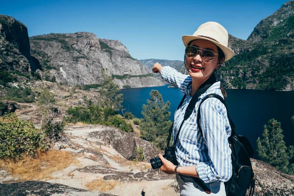 Genç kız Asya backpacker yosemite Milli Parkı ziyaret edin. zarif kadın gezgin işaret mavi gökyüzü ve temiz Gölü yüz kamera gülümseyen güneş gözlüklü. seyahat san franisco yaşam tarzı kavramı.