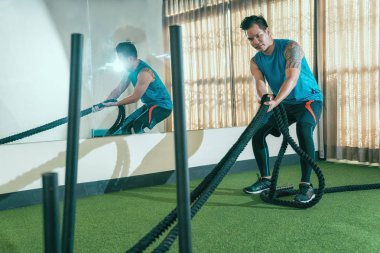 Sağlık kulübünde oda kadar düzenli ağır savaş ip çekerek yorgun kaslı erkek eğitmen. Genç çalıştırıcı mücadele tutan spor salonunda ipleri. yakışıklı Asya spor giyim ve silah dövme ile.