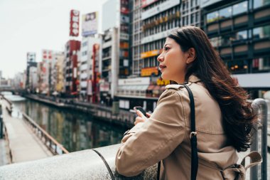 Genç Asyalı kadın avukat gezi osaka City otel onun iş seyahat etmek içinde arama mobil akıllı telefon tutarak etrafa bakıyor. şehirde kadın profesyonel Köprü Nehri üzerinde durmak.