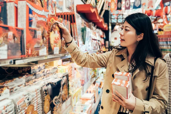 asian girl traveler in her business trip visiting local specialty shop in dotonbori osaka japan choosing souvenir for family. young lady looking holding the japanese cookie in the store.